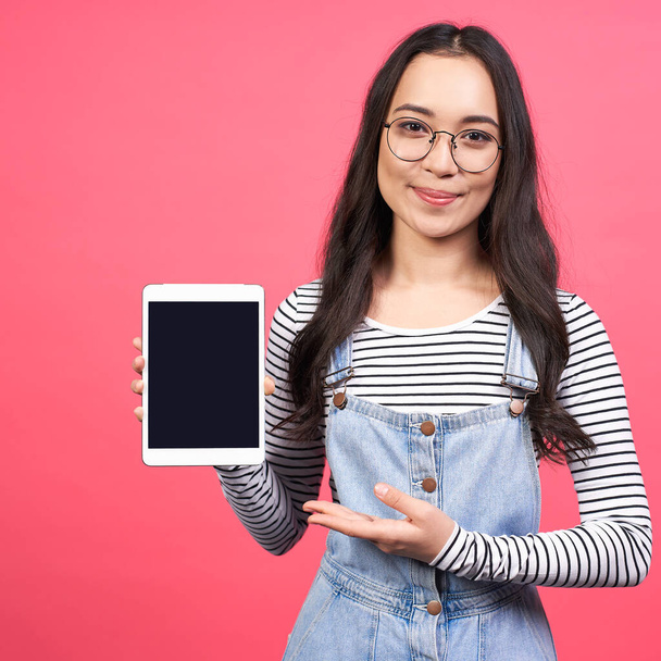 The best choice. Cute young Asian girl in glasses and striped jacket shows her hand on the tablet, stands against a pink background. The concept of people and technology, modern design. - Fotoğraf, Görsel