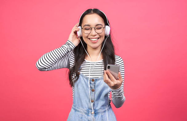 Happy asian girl relaxed with a great song, with a wide smile, touching headphones, listening to music, wearing a striped sweater and denim sundress, isolated on a pink studio wall, wants to dance. - Photo, image