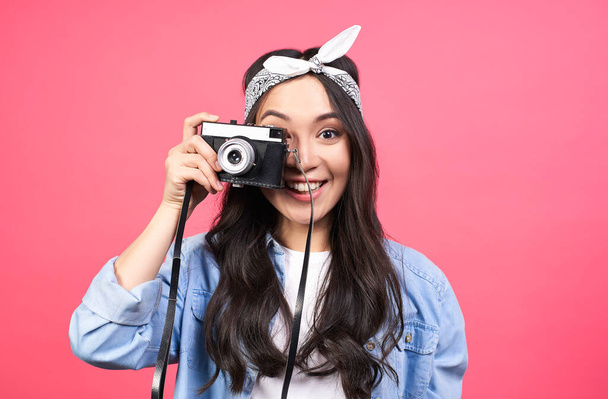 Portrait of a beautiful young dark-haired Asian woman, in casual stylish denim clothes, standing isolated on a pink background, taking pictures with a retro vintage camera. - Foto, Bild