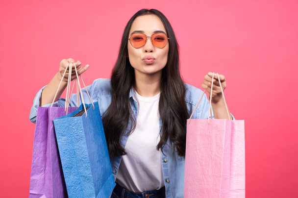 Portrait of an excited beautiful girl in a white T-shirt and denim shirt and red sunglasses, holding multi-colored bags, packages, isolated on a pink background. - Foto, Bild