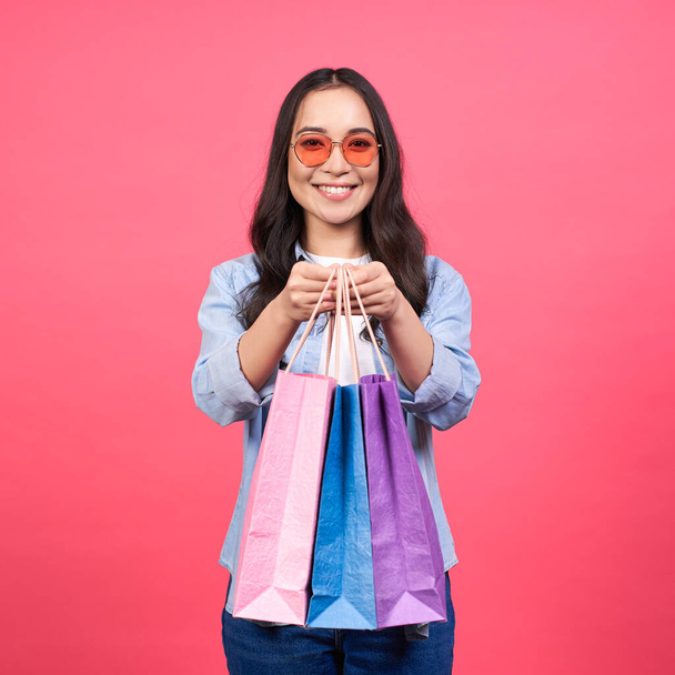 Happy tender young Asian woman with long hair, in denim clothes and bright glasses, holds many multi-colored paper bags in her hands, cheerful after shopping, holds out the bags to the camera. - Fotografie, Obrázek