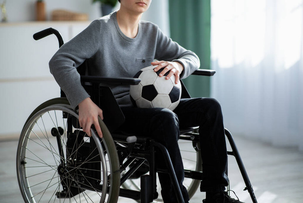 車椅子に座っているサッカーボールを持つ障害者の少年の作物のビュー,自宅で彼の負傷に動揺を感じます - 写真・画像