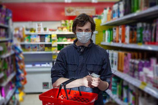 Man wears disposable face mask, makes shopping, thinks about lack of money during coronavirus pneumonia outbreak, poses with basket in department store. Concept of a crisis during an epidemic - Fotó, kép
