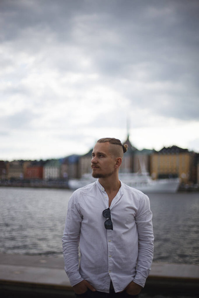 Ein junger Mann mit Schnurrbart und Bart im weißen Hemd steht am Ufer eines Sees oder Flusses vor dem Hintergrund der Altstadt. schaut der Mann zur Seite und hält seine Hände in den Taschen. bewölkter Tag - Foto, Bild