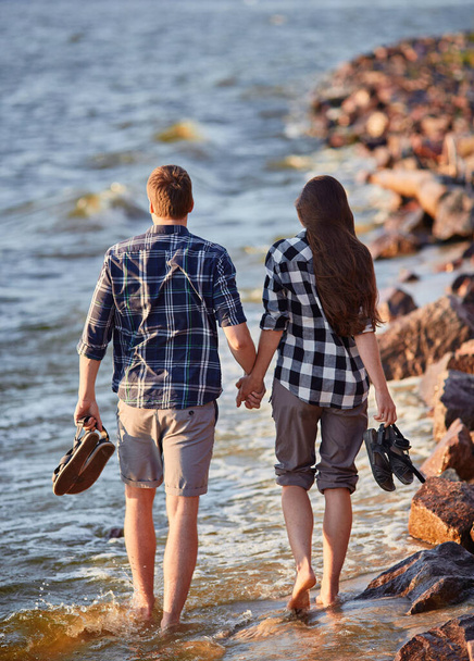 Happy caucasian fashionable couple in love holding hands and walking on coast near river. In background is sunset. - Photo, Image
