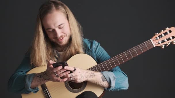 Jonge langharige blonde man met gitaar sms 'en met vrienden op smartphone tijdens de repetitie over zwarte achtergrond. Muziekconcept. Moderne technologie - Video
