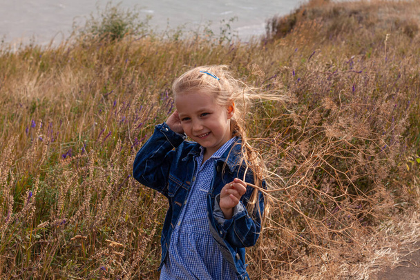 Adorable little girl in denim jacket back view on a hill with sea landscape view. Stylish child with long blonde hair on countryside cloudy sky background. Outdoor walking rural road trip faceless. - Photo, Image
