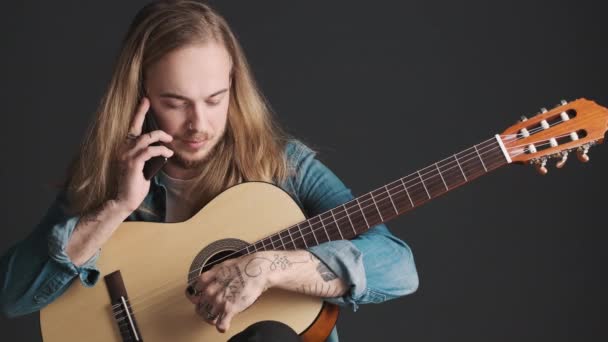 Fiatal szakállas férfi zenész gitárral beszél okostelefonon próba közben a stúdióban elszigetelt fekete háttérrel. Zenei koncepció - Felvétel, videó
