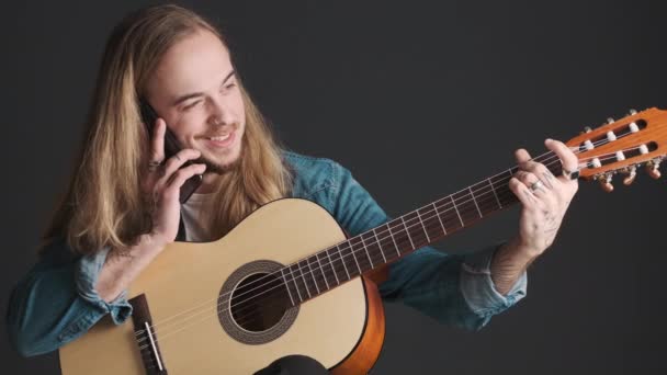 Jovem músico de cabelos compridos falando no smartphone enquanto ajusta a guitarra sobre fundo preto. Conceito musical - Filmagem, Vídeo
