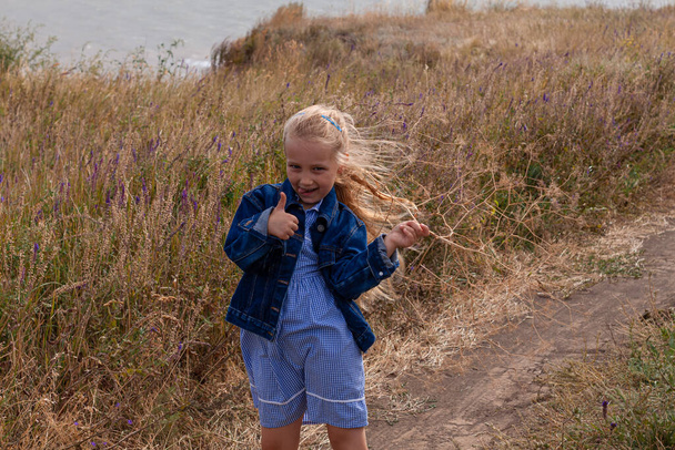 Чарівна маленька дівчинка в джинсовій куртці вид ззаду на пагорб з видом на море. Стильна дитина з довгим світлим волоссям на сільському фоні хмарного неба. Відкрита прогулянка по сільській дорозі безлика
. - Фото, зображення