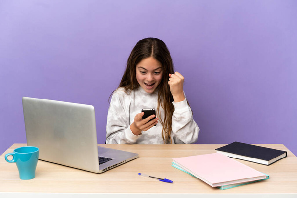 Mała uczennica w miejscu pracy z laptopem odizolowanym na fioletowym tle zaskoczona i wysyłająca wiadomość - Zdjęcie, obraz
