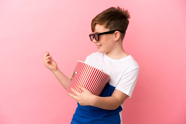 Little redhead boy isolated on pink background with 3d glasses and holding a big bucket of popcorns while looking side - Foto, Bild