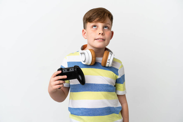Ένα μικρό κοκκινομάλλικο αγόρι που κρατάει ένα παιχνίδι απομονωμένο σε λευκό φόντο σκεπτόμενο μια ιδέα ενώ κοιτάζει ψηλά - Φωτογραφία, εικόνα