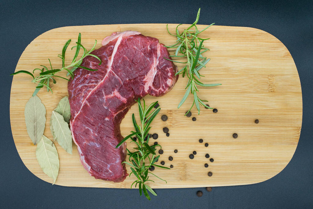 Stek wołowy, widok świeżej wołowiny na drewnianej desce z rozmarynem, czarny pieprz, liść toalety. - Zdjęcie, obraz