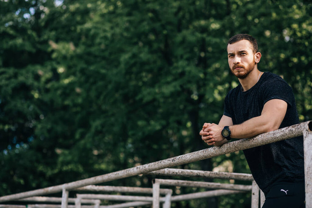 Portrait young bearded man standing on public sports ground and training on parallel bars. Handsome workout athlete. Portrait sporty man with beard, rest after training. - Valokuva, kuva