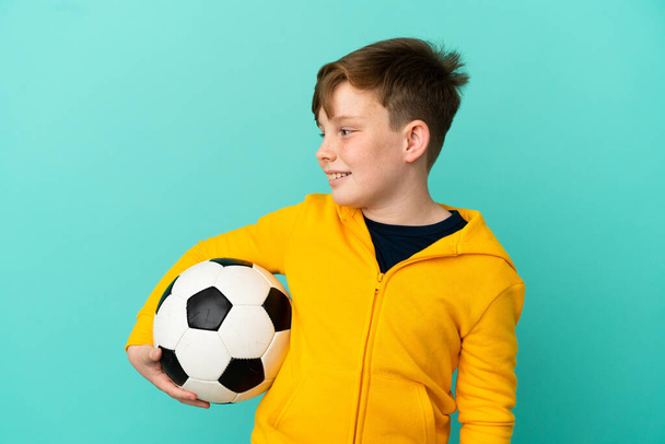 Redhead dítě hraje fotbal izolované na modrém pozadí při pohledu do strany a usmívá se - Fotografie, Obrázek