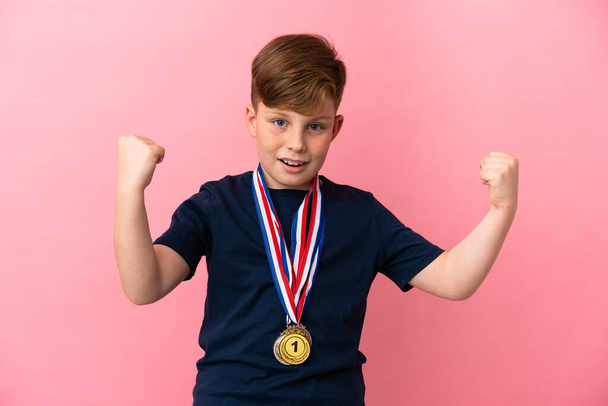 Malý ryšavý chlapec s medailemi izolované na růžovém pozadí slaví vítězství - Fotografie, Obrázek