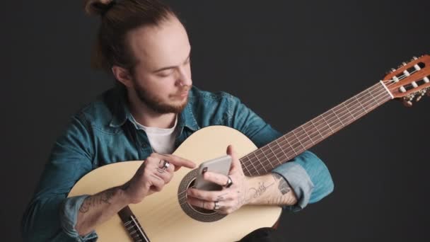 Jonge stijlvolle bebaarde man met gitaar controleren van sociale netwerken op smartphone wachten op repetitie te beginnen over zwarte achtergrond - Video