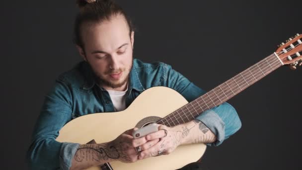 Jonge stijlvolle bebaarde man op zoek gelukkig vinden hoe te stemmen gitaar met behulp van smartphone voor het tijdens de repetitie in de studio geïsoleerd op zwarte achtergrond - Video
