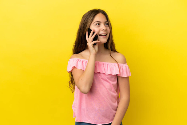 Little caucasian girl isolated on yellow background keeping a conversation with the mobile phone - Fotografie, Obrázek