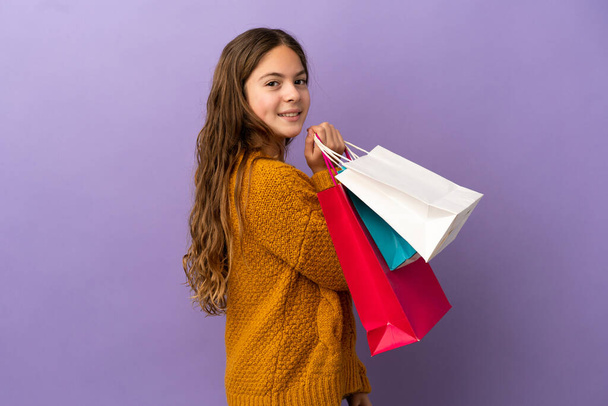 Pequeña chica caucásica aislada sobre fondo púrpura sosteniendo bolsas de compras y sonriendo - Foto, imagen