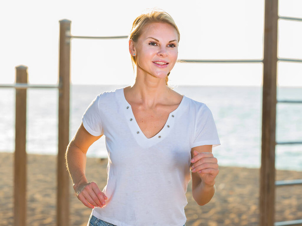 Smiling adult woman in white T-shirt is jogging on the beach - Valokuva, kuva