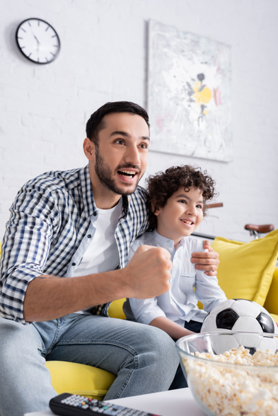 podekscytowany arabski człowiek pokazując gest sukcesu podczas oglądania meczu piłki nożnej wraz z synem w domu - Zdjęcie, obraz
