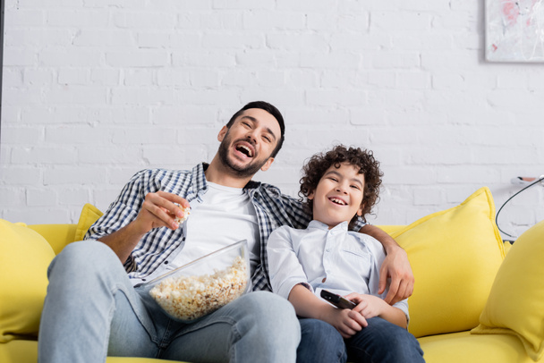 śmiejący się muzułmanin jedzący popcorn podczas oglądania filmu z synem w domu - Zdjęcie, obraz