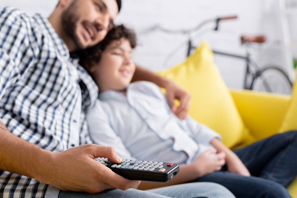 selective focus of tv remote controller in hand of muslim man watching movie with son, blurred background - Photo, Image