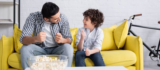 feliz chico árabe mostrando el pulgar hacia arriba mientras habla con el padre en el sofá en casa, pancarta - Foto, imagen