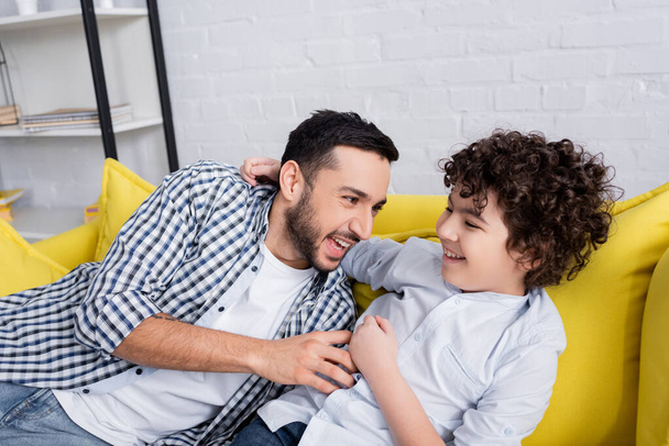 aufgeregter muslimischer Mann kitzelt glücklichen Sohn, während er Spaß auf dem Sofa hat - Foto, Bild