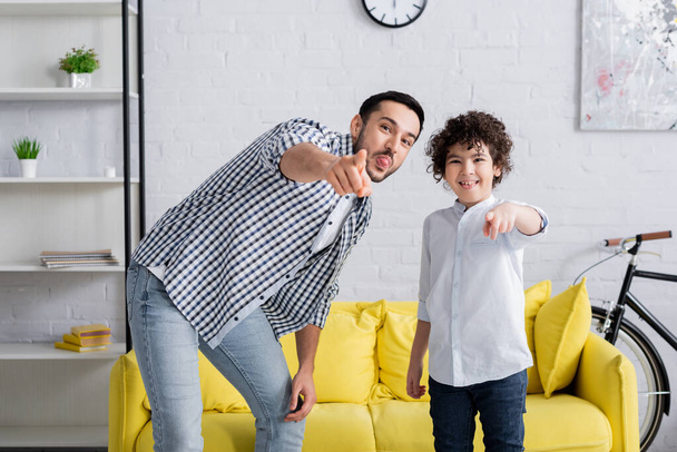 cheerful arabian man and son sticking tongues and pointing with fingers while looking at camera - Photo, Image