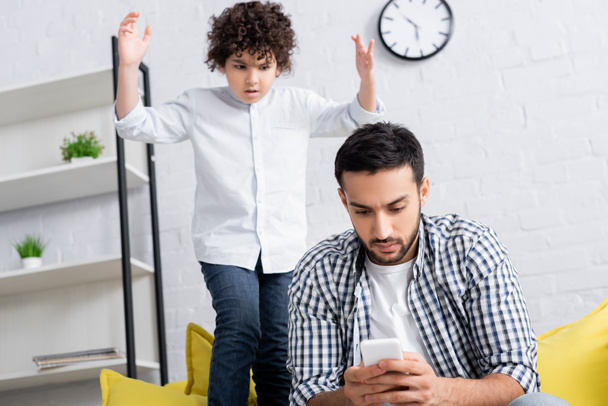 displeased arabian boy standing with raised hands near serious father messaging on smartphone - Photo, Image
