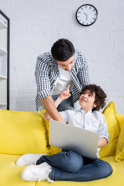 smiling arabian kid sitting on sofa with laptop and pointing with finger near father - Photo, Image