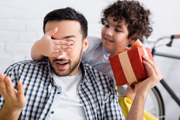 muslim boy covering eyes of father holding gift box, blurred background - Photo, Image