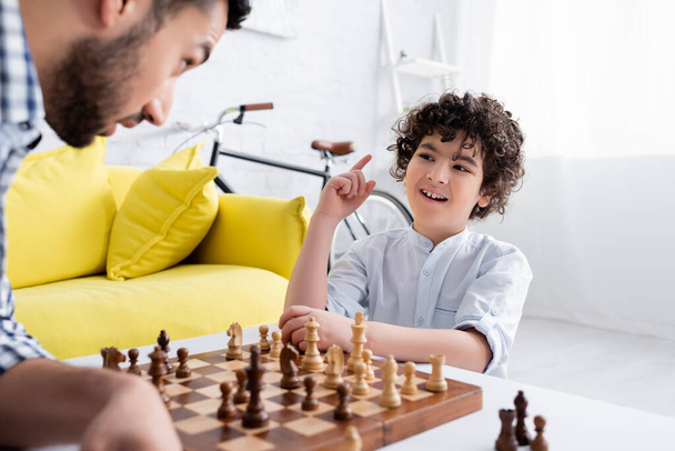 alegre mulsim chico señalando con el dedo mientras juega al ajedrez con padre en primer plano borrosa - Foto, Imagen