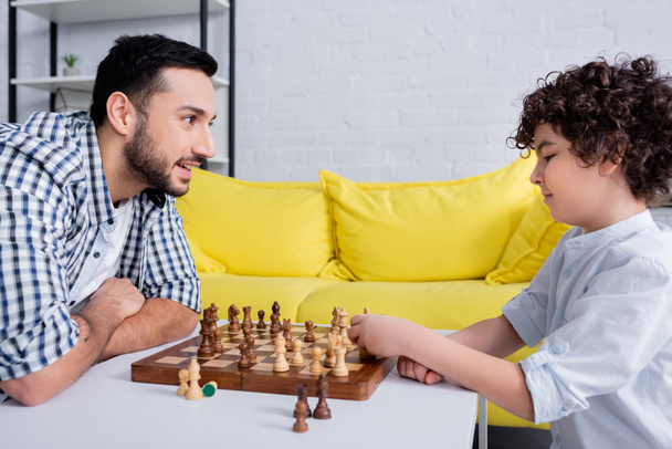 Lächelnder muslimischer Mann sieht Sohn zu Hause Schach spielen  - Foto, Bild