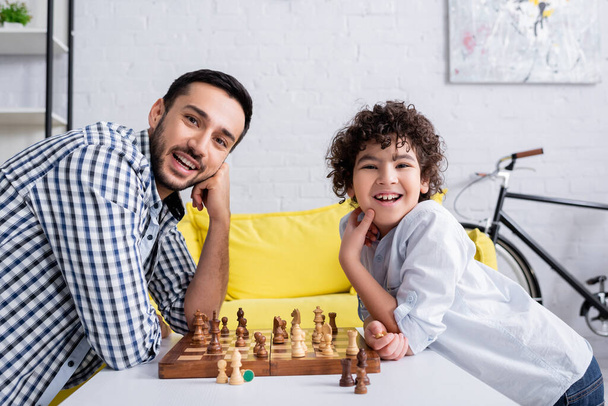 Fröhlicher arabischer Vater und Sohn blicken in die Kamera beim Schach auf dem Tisch  - Foto, Bild