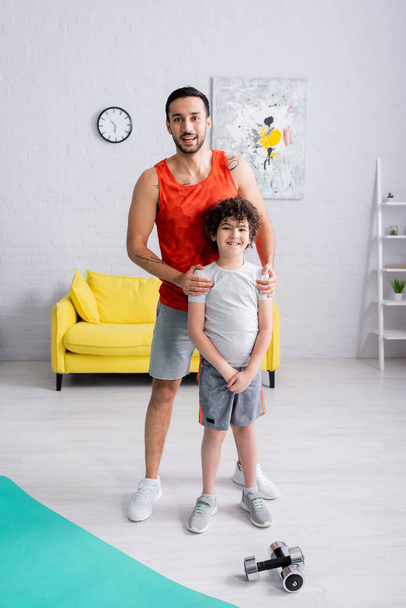 Glimlachende Arabische man knuffelende zoon in de buurt halters en fitness mat thuis  - Foto, afbeelding