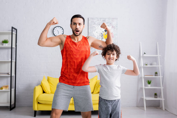 Lächelnder arabischer Junge zeigt Muskeln neben Vater in Sportbekleidung  - Foto, Bild
