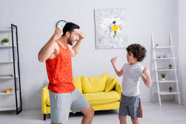 Emocionado pai muçulmano e filho mostrando músculos na sala de estar  - Foto, Imagem