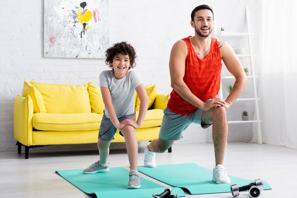 Smiling muslim son and father doing lunges near dumbbells at home  - Photo, Image