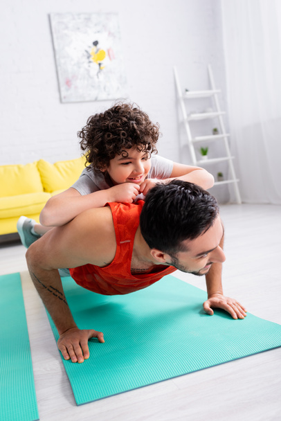 Sonriente chico árabe en la espalda del padre de entrenamiento en la alfombra de fitness en casa  - Foto, imagen