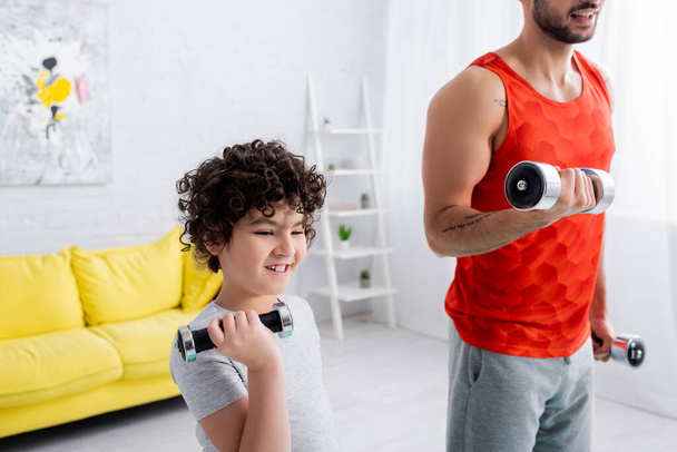 Smiling muslim kid exercising with dumbbell near father at home  - Photo, Image