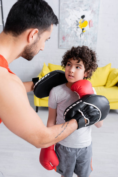 Muslim father in punch mitts teaching son in boxing gloves at home  - Photo, Image