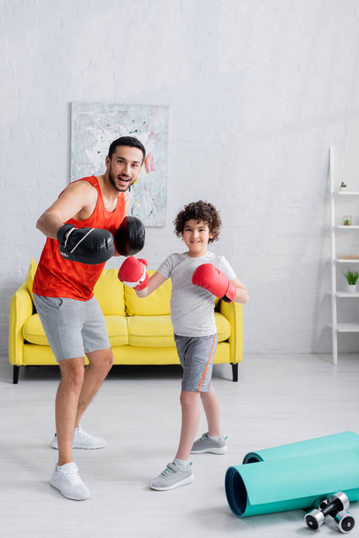 Smiling arabian father and son in boxing gloves smiling at camera near sport equipment at home  - Photo, Image