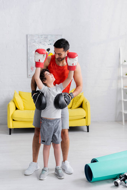 Muslim boy in boxing gloves looking at father near fitness mats and dumbbells  - Photo, Image