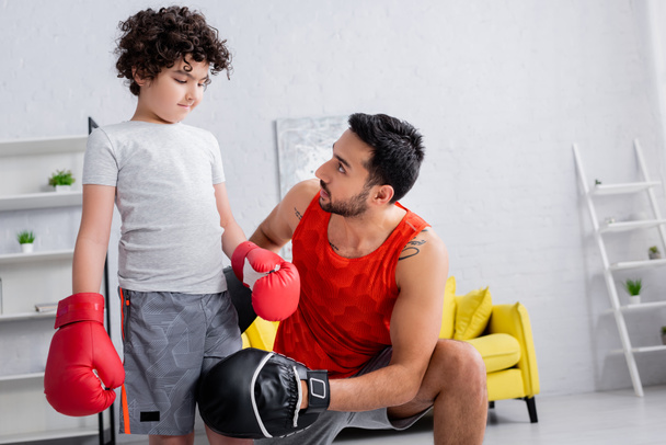 Muçulmano em punch mitts de pé perto do filho em luvas de boxe na sala de estar  - Foto, Imagem