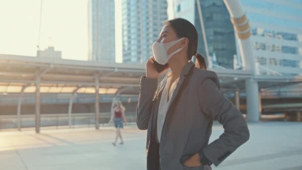 Moda ofisindeki genç Asyalı iş kadını şehir dışında tek başına yürürken telefonla tıbbi maske takıyor. COVID-19 kavramının yayılmasını önlemek için sosyal mesafe. - Video, Çekim