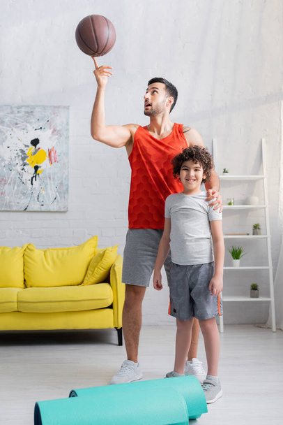 Arabo padre tenendo pallacanestro sul dito vicino allegro figlio e stuoie fitness  - Foto, immagini